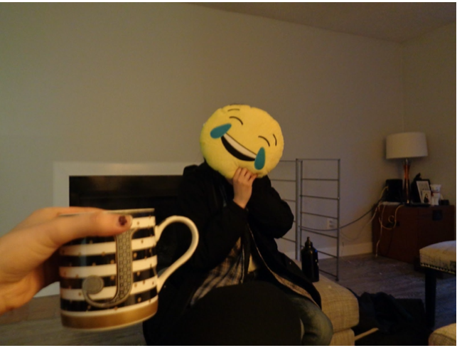 person with a black coat holds a laughing emoji over their face. A hand holds a mug out with the letter J on it
